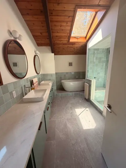 Skylit bathroom finished in sage green tile with a freestanding tub and glass shower