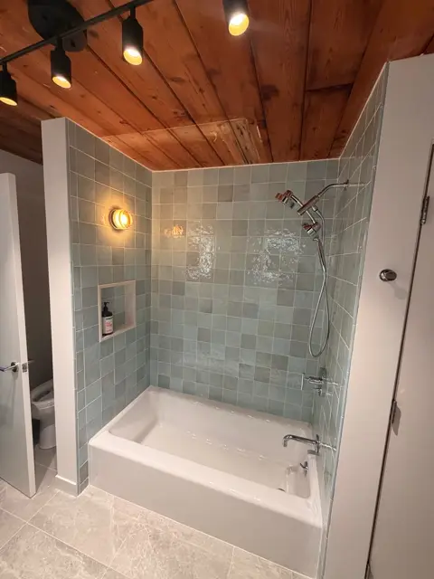 Tub and shower combination finished in sage green tile under a wood-beamed ceiling